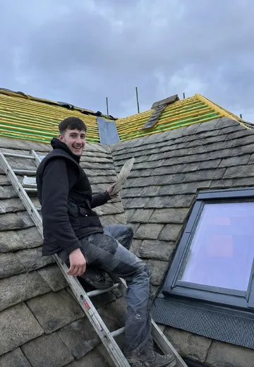 Adam sitting on a roof ladder over a Cotswold stone slate roof, holding a slate, with the slope above him stripped back to battens.