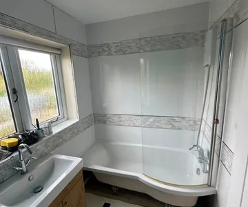 A bathroom with a shower bath and a glass screen, tiled walls with a mosaic border running through them, and a basin set on a timber vanity unit.