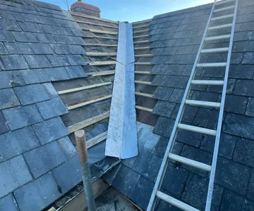 A slate roof with the valley stripped back to new timber battens and a new lead valley dressed down the middle of it, with a ladder up the slope alongside.