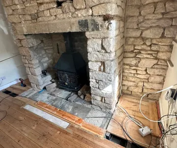 A stone inglenook fireplace with a cast iron stove standing in the opening, the chimney breast and wall stripped back to bare stone, and a section of floorboards lifted in front of it.