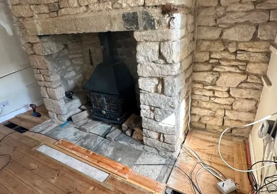 A stone inglenook with a cast iron stove in the opening, the chimney breast back to bare stone and the floor up in front of it.